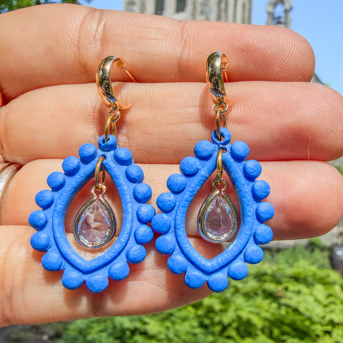 Boucles d’oreilles Qatra – Gouttes de lumière : pendentifs bleu cobalt bordés de perles d’argile et gouttes irisées, tenus dans la main