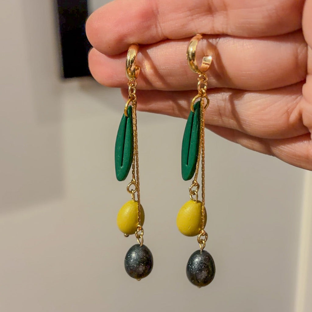 Paire de boucles d’oreilles à olives verte et noire, feuilles allongées et chaînes dorées, présentée dans une main.