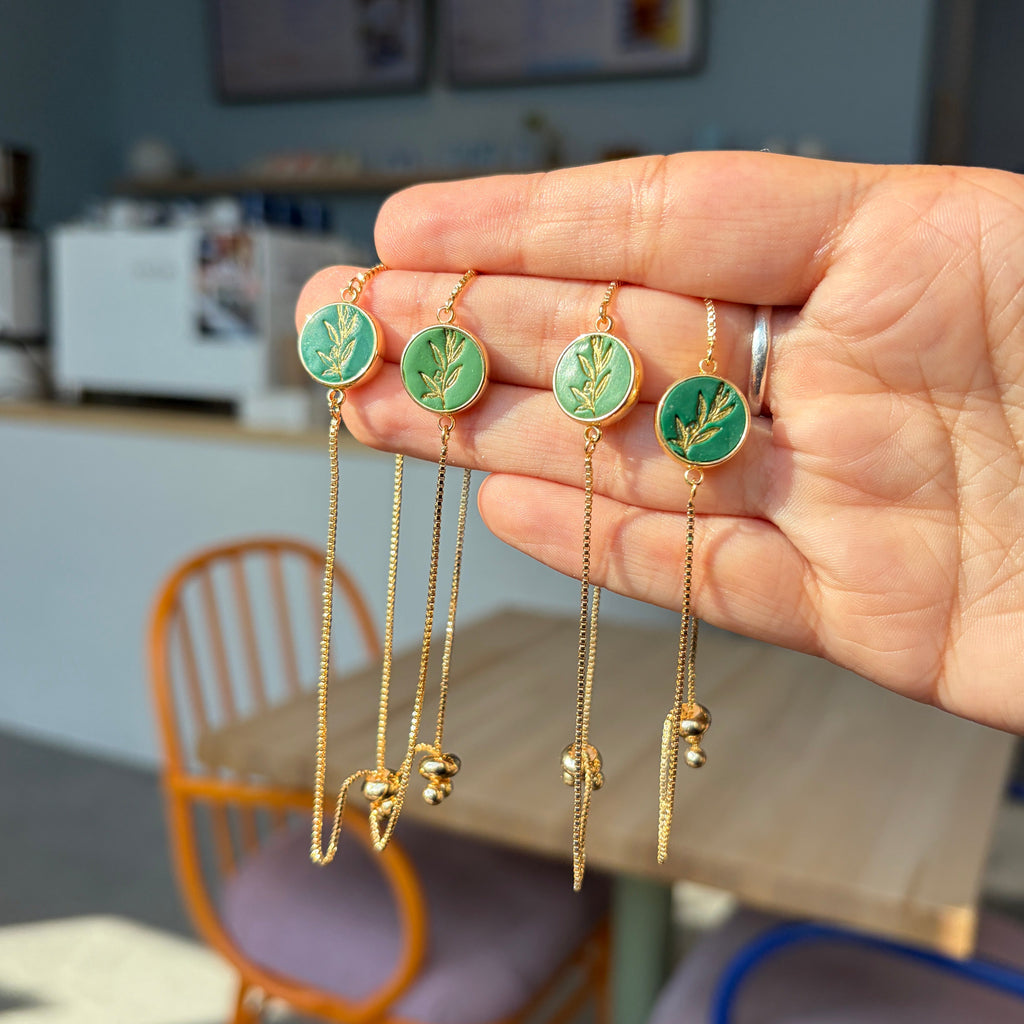 Quatre bracelets Olea à médaillon rond vert, motif de rameau d’olivier doré et fine chaîne dorée, présentés dans une main.