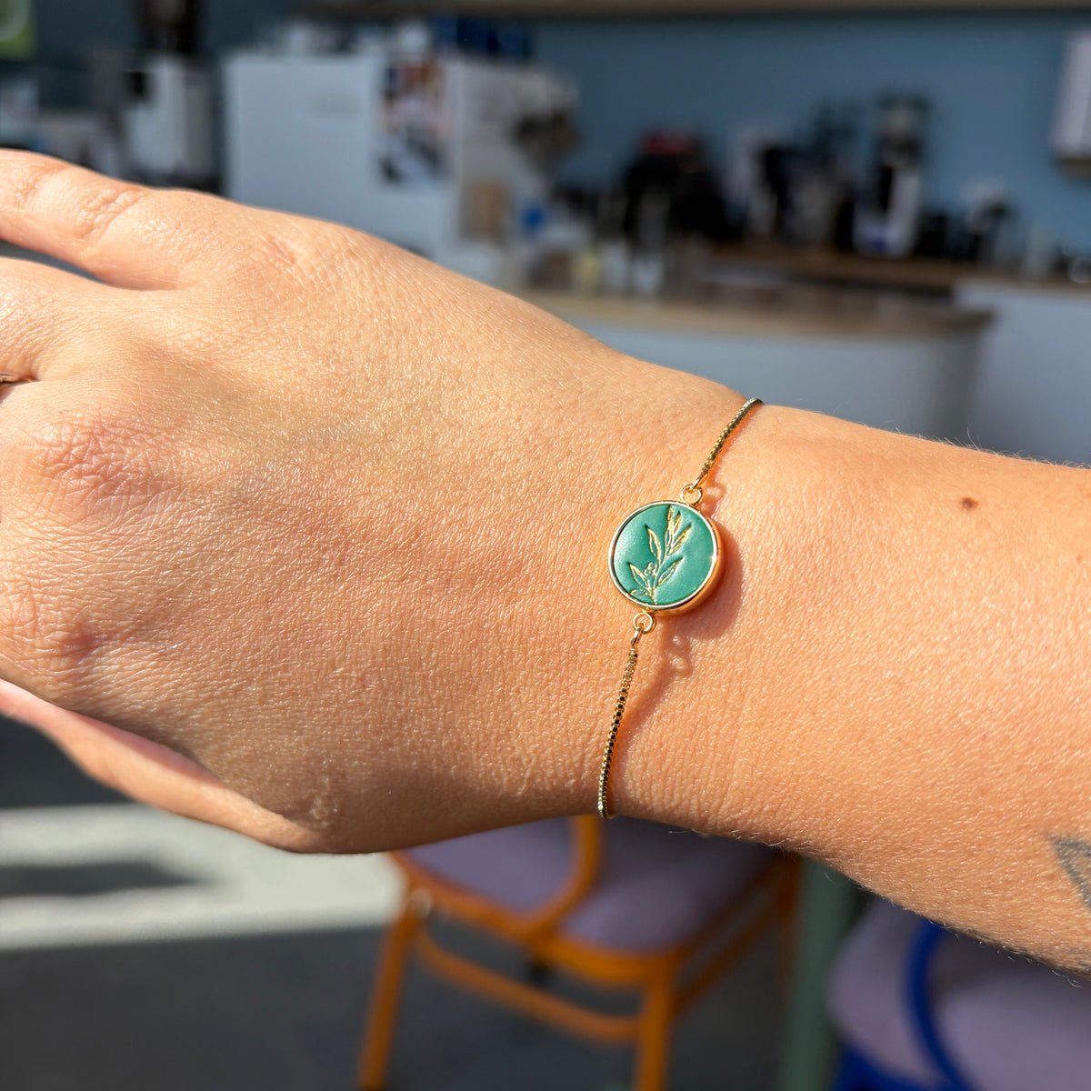 Bracelet Olea porté au poignet : médaillon rond vert orné d’un rameau d’olivier doré et fine chaîne de couleur dorée.