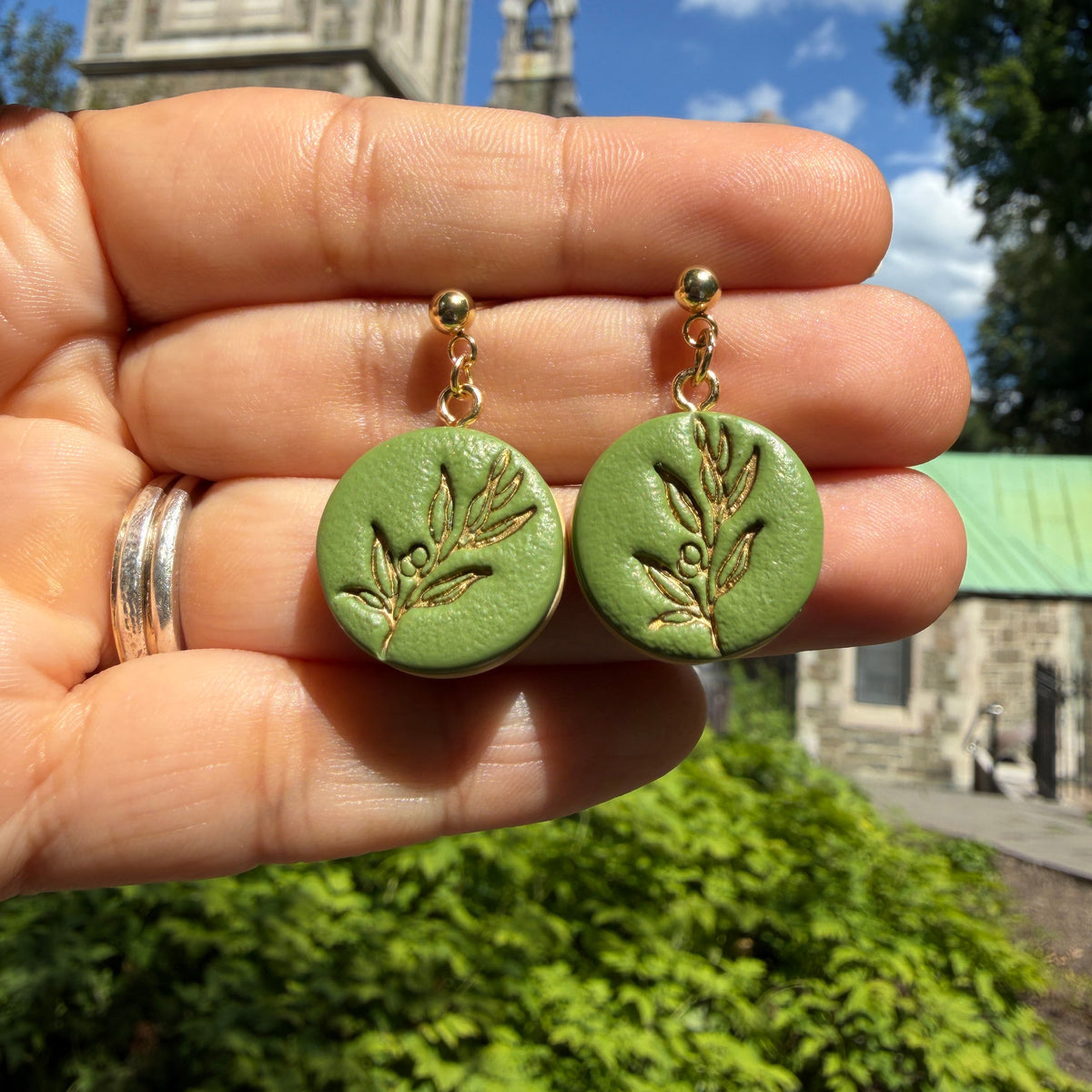 Boucles d’oreilles Olea – Murmure d’olivier : médaillons ronds verts à rameau d’olivier rehaussé de doré, présentés dans la main.