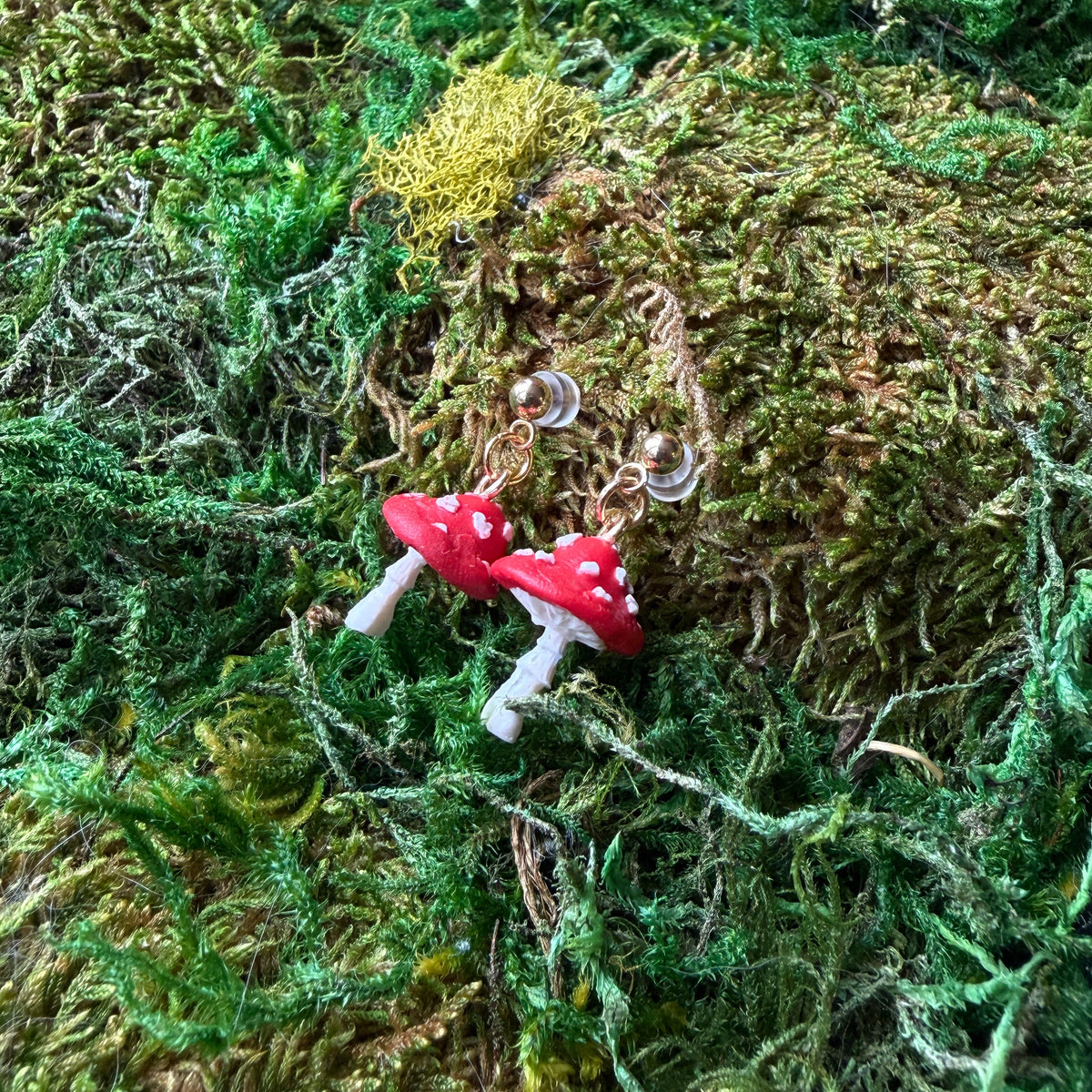 Paire de boucles d’oreilles amanites en argile polymère rouge et blanche, posée sur la mousse.