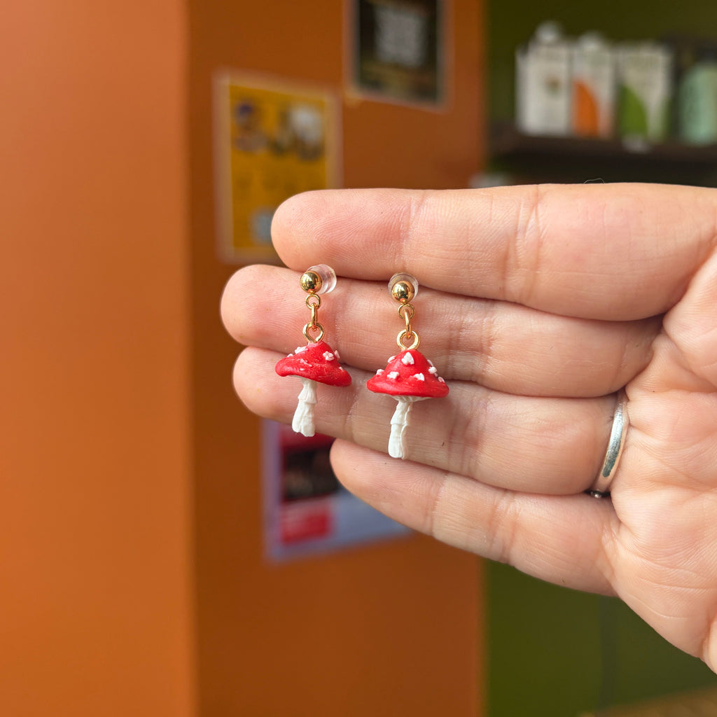 Boucles d’oreilles pendantes avec champignons rouges à pois blancs et clous dorés, présentées dans une main.
