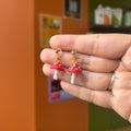 Boucles d’oreilles pendantes avec champignons rouges à pois blancs et clous dorés, présentées dans une main.
