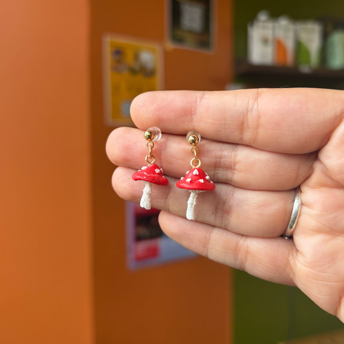 Boucles d’oreilles pendantes avec champignons rouges à pois blancs et clous dorés, présentées dans une main.