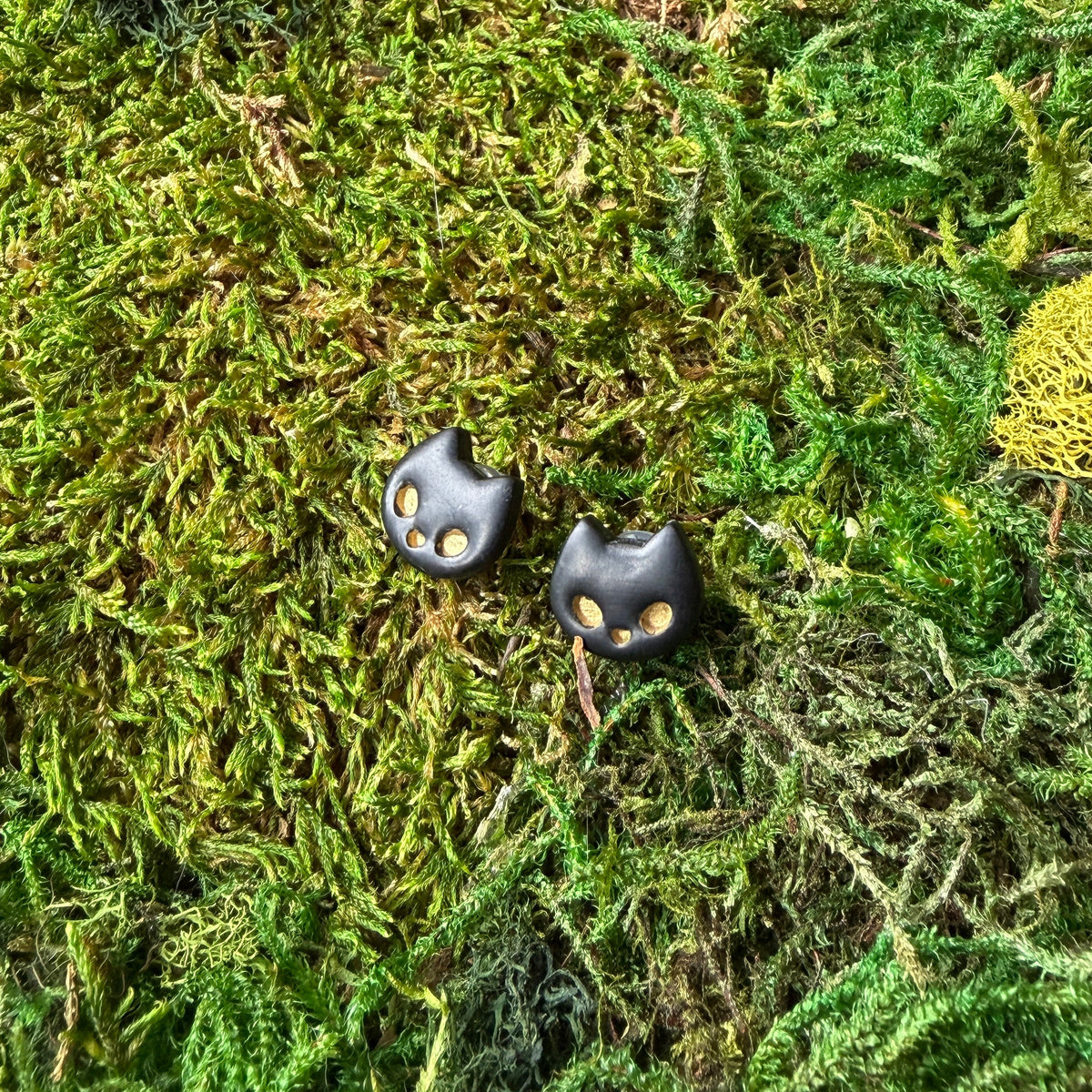 Deux puces chats noirs en argile polymère avec yeux dorés, posées sur la mousse.
