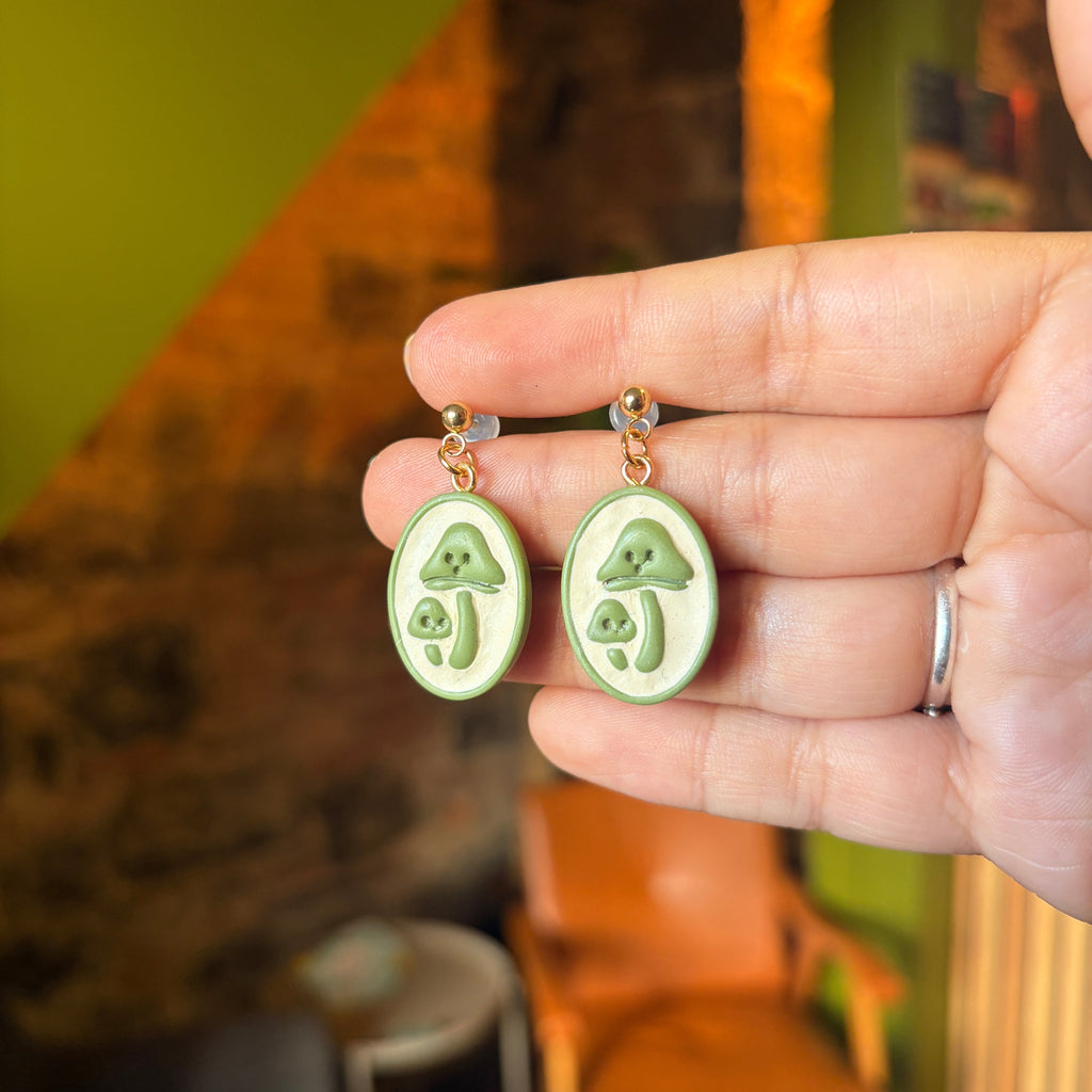 Boucles d’oreilles Rêverie des sous-bois : champignons beiges en relief sur médaillons verts et clous boule dorés, présentés dans une main.