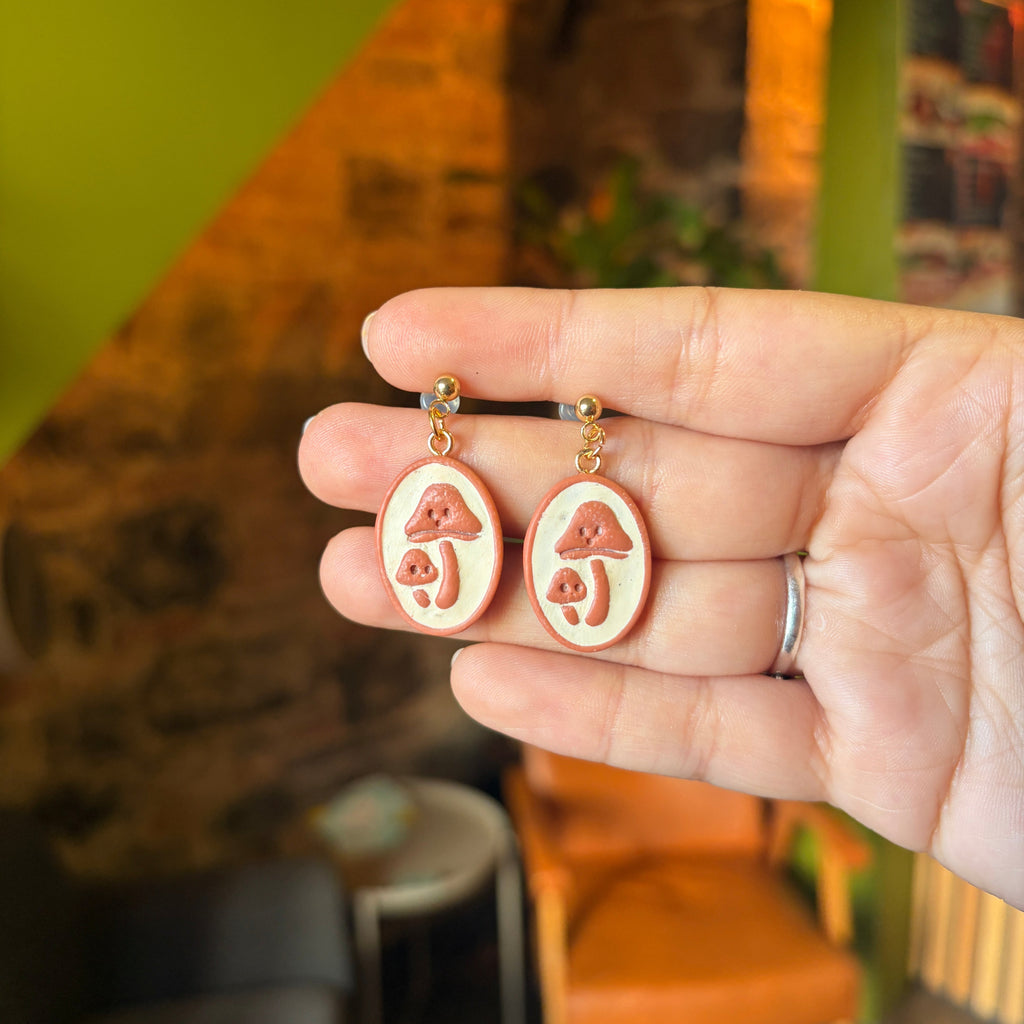 Boucles d’oreilles Rêverie des sous-bois : champignons terracotta en relief sur médaillons beiges et clous boule dorés, présentés dans une main.