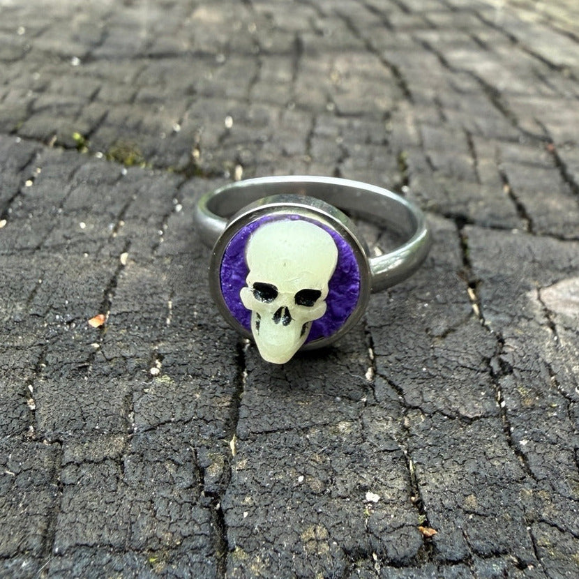 Bague à crâne sculpté ivoire et noir sur fond violet, avec monture argentée.