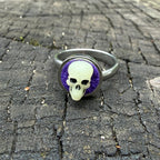 Bague à crâne sculpté ivoire et noir sur fond violet, avec monture argentée.