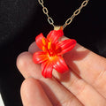 Pendentif en forme de fleur rouge et orange à cœur et étamines jaunes, porté sur une chaîne dorée.