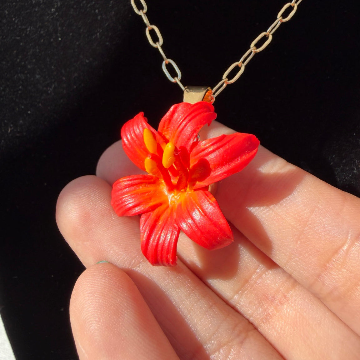 Pendentif en forme de fleur rouge et orange à cœur et étamines jaunes, porté sur une chaîne dorée.