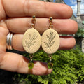 Boucles d’oreilles Olea – Rameau d’or : ovales beige à rameau d’olivier noir et gouttes de verre vert, tenus dans la main