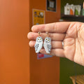 Boucles d’oreilles Veilleurs de lune : hiboux blancs aux yeux jaunes et au plumage souligné de noir, suspendus à des clous sphériques de couleur dorée.